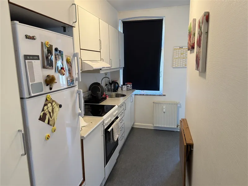 En køkken med hvide vægge og gulv, hvide skabe, en køleskab og en ovn og en kalender på væggen