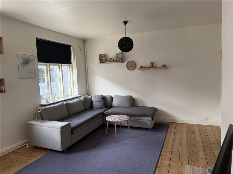 Lyst og moderne stue med lysegule vægge og trægulv. Komforabel grå sofa med pudsede høje hovedstøtter. Rundt træbord med