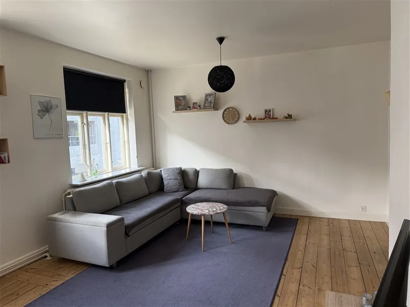 Lyst og moderne stue med lysegule vægge og trægulv. Komforabel grå sofa med pudsede høje hovedstøtter. Rundt træbord med