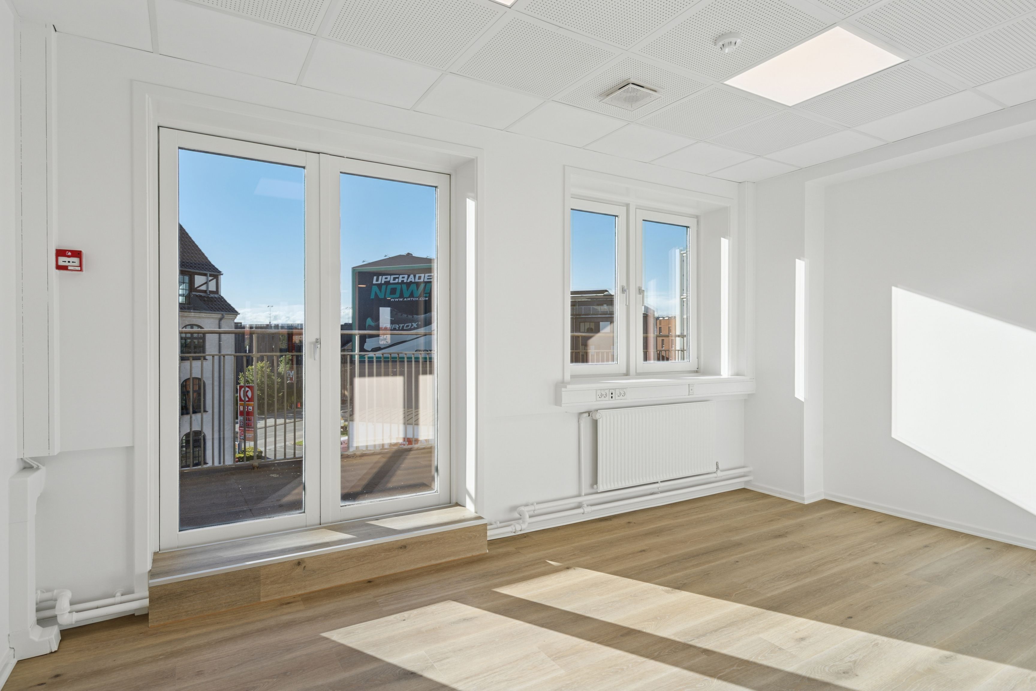 Et moderne kontor med lyse gulve, hvide vægge, vinduer og en terrasse med udsigt over byen. Værelset har to vinduer, hvoraf