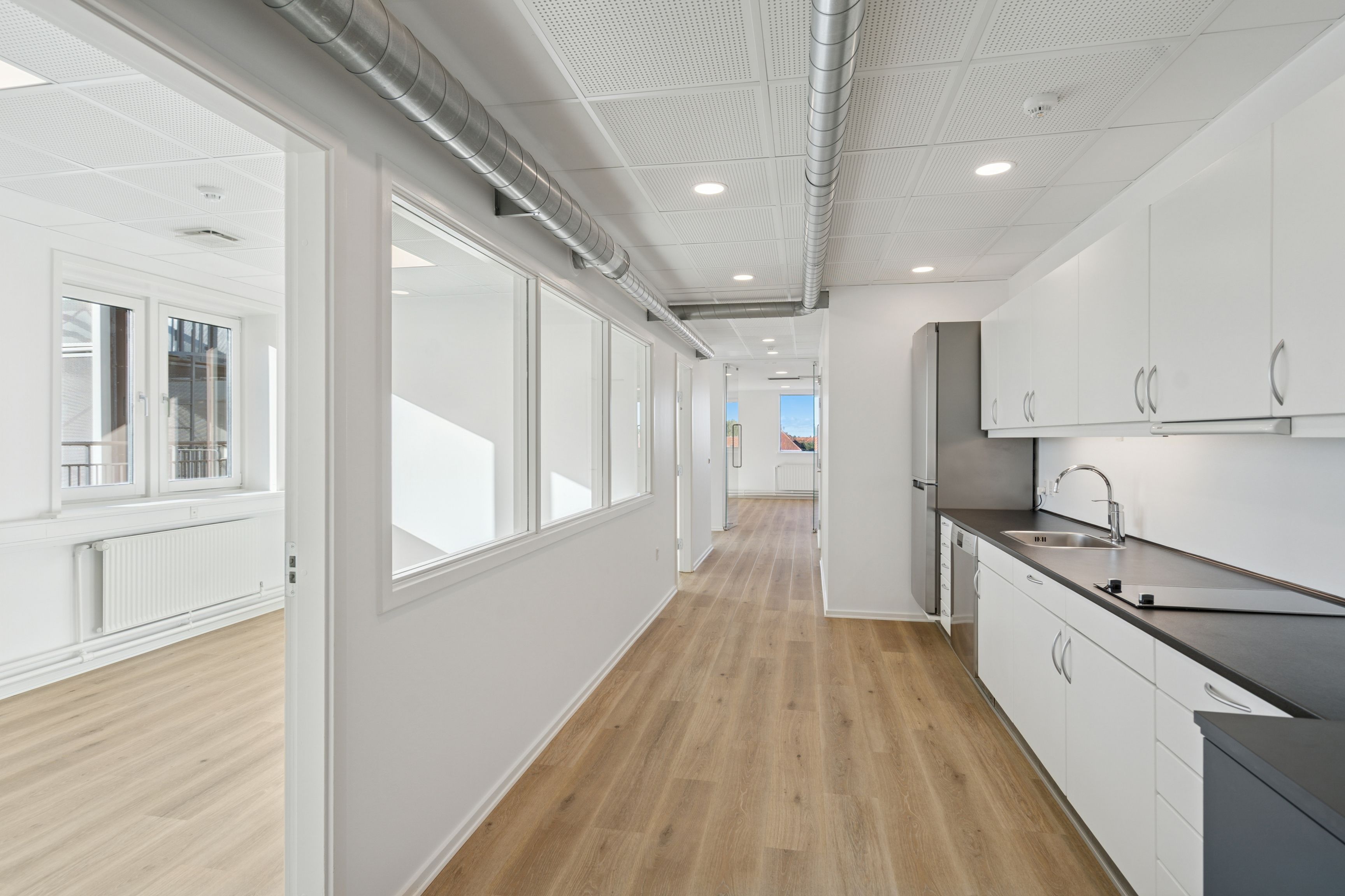 Kontor med hvidt loft, hvid væg, trægulv og moderne møbler. Køkken med sort kammer, hvidt loft og moderne køkkenindretning.