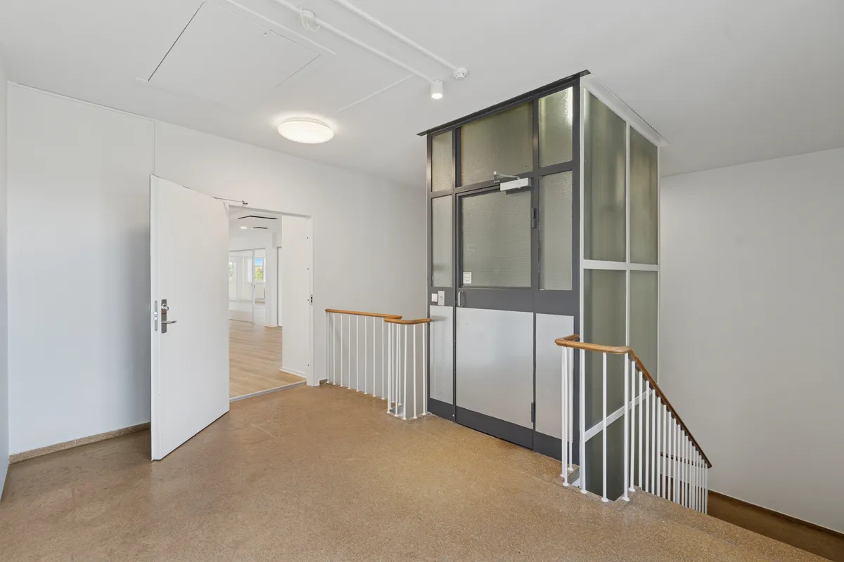 Et elegant og moderne kontorlokal med lyse lofter og hvidvaskede vægge. En slank elevator med glasdøre og et moderne