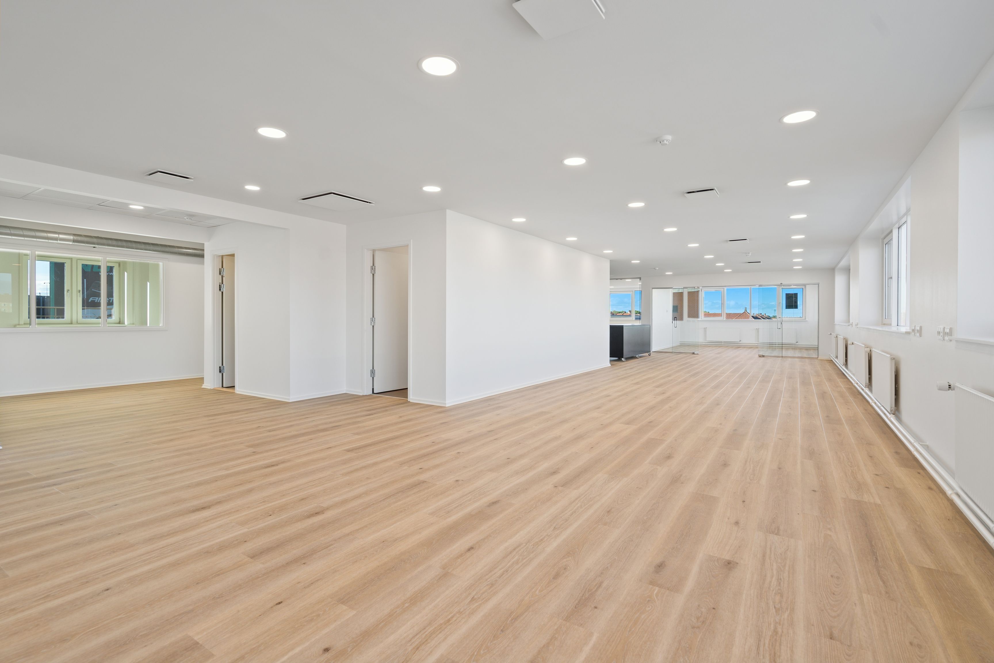 En stor, tom kontorhal med hvide vægge og lyse trægulve samt vinduer med udsigt til byen.