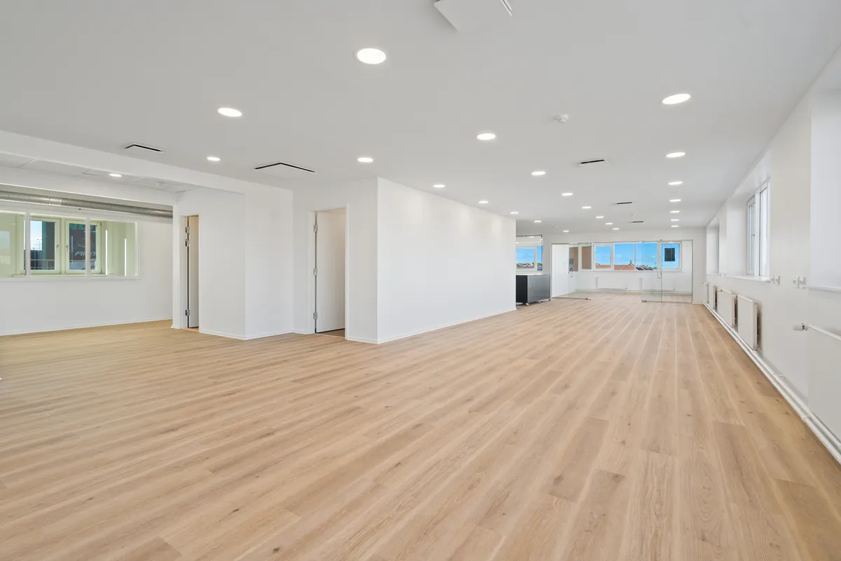 En stor, tom kontorhal med hvide vægge og lyse trægulve samt vinduer med udsigt til byen.