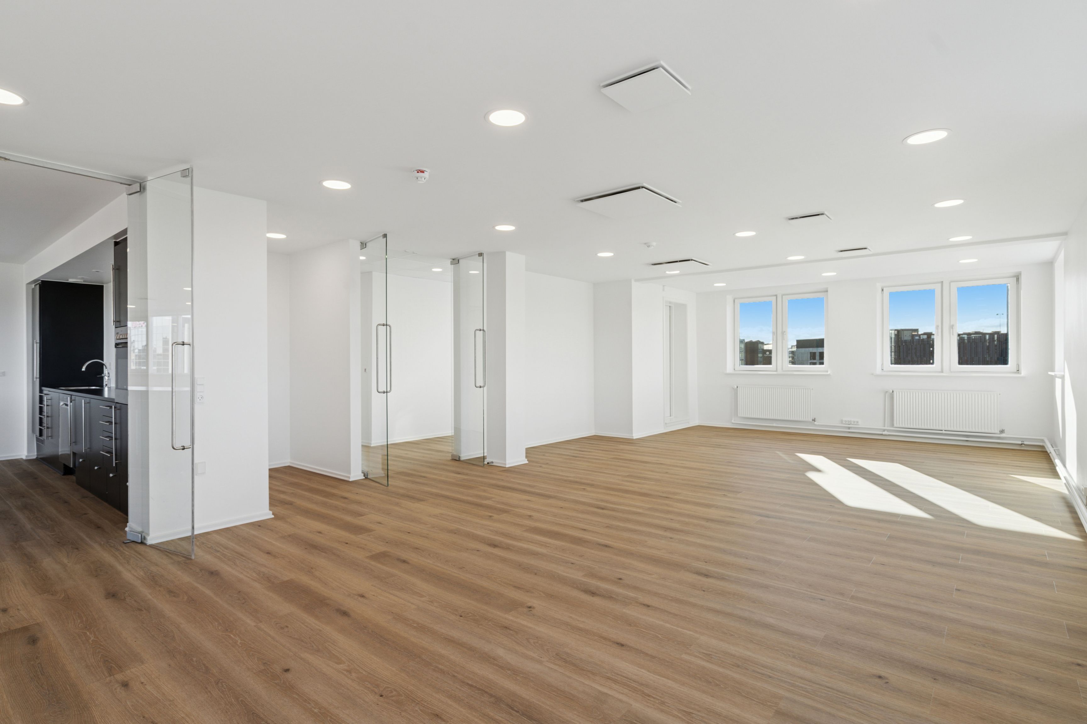 Kontor med højloft og glasdøre, hvide vægge og trægulv, tre store vinduer med udsigt, radiatorer og tæppe på gulvet, hvidt