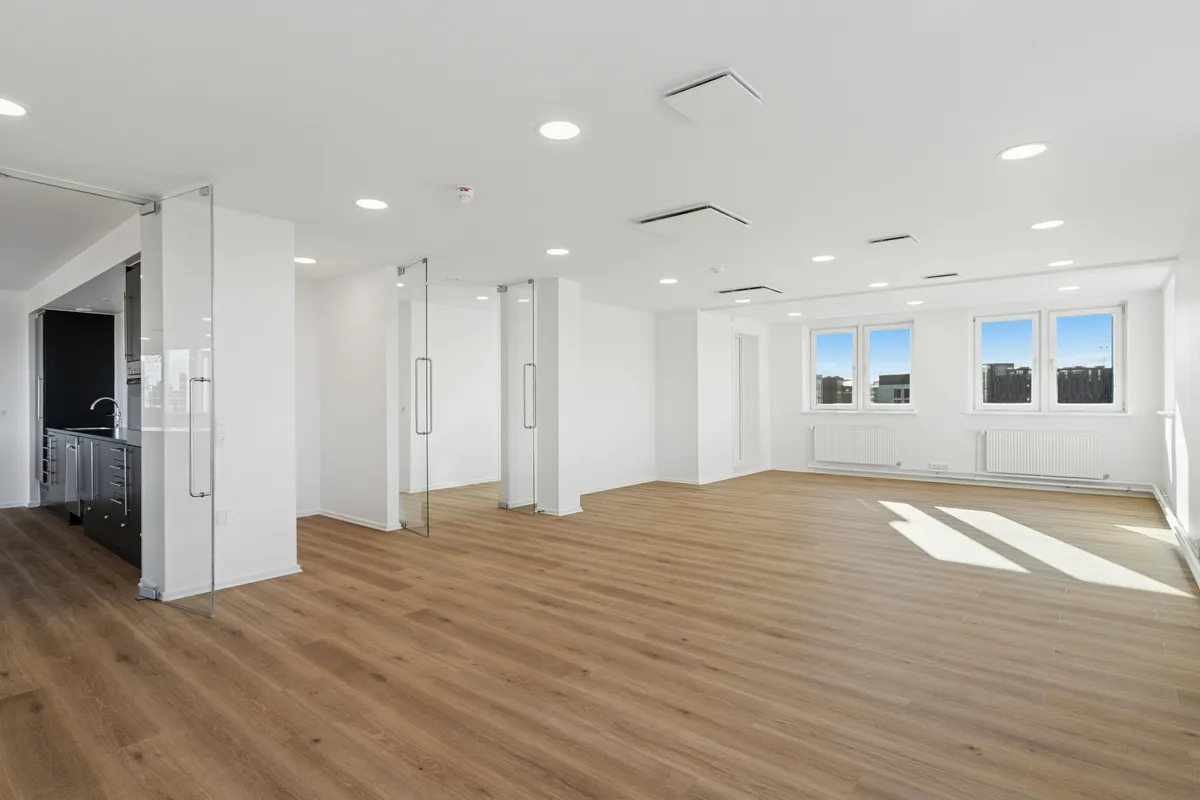 Kontor med højloft og glasdøre, hvide vægge og trægulv, tre store vinduer med udsigt, radiatorer og tæppe på gulvet, hvidt