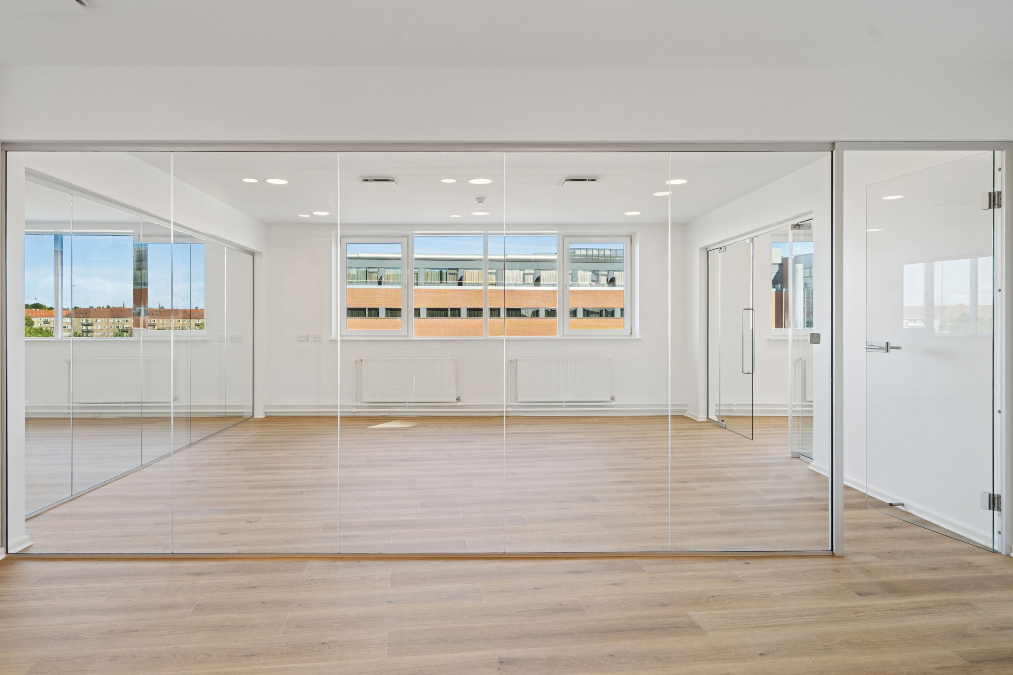 En tom kontormængde med store glasvægge, lyse loftsbelægninger og en tagterrasse med udsigt til bygninger og grønne områder