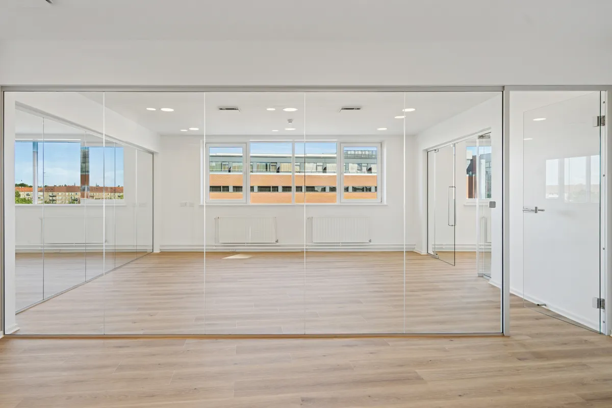 En tom kontormængde med store glasvægge, lyse loftsbelægninger og en tagterrasse med udsigt til bygninger og grønne områder