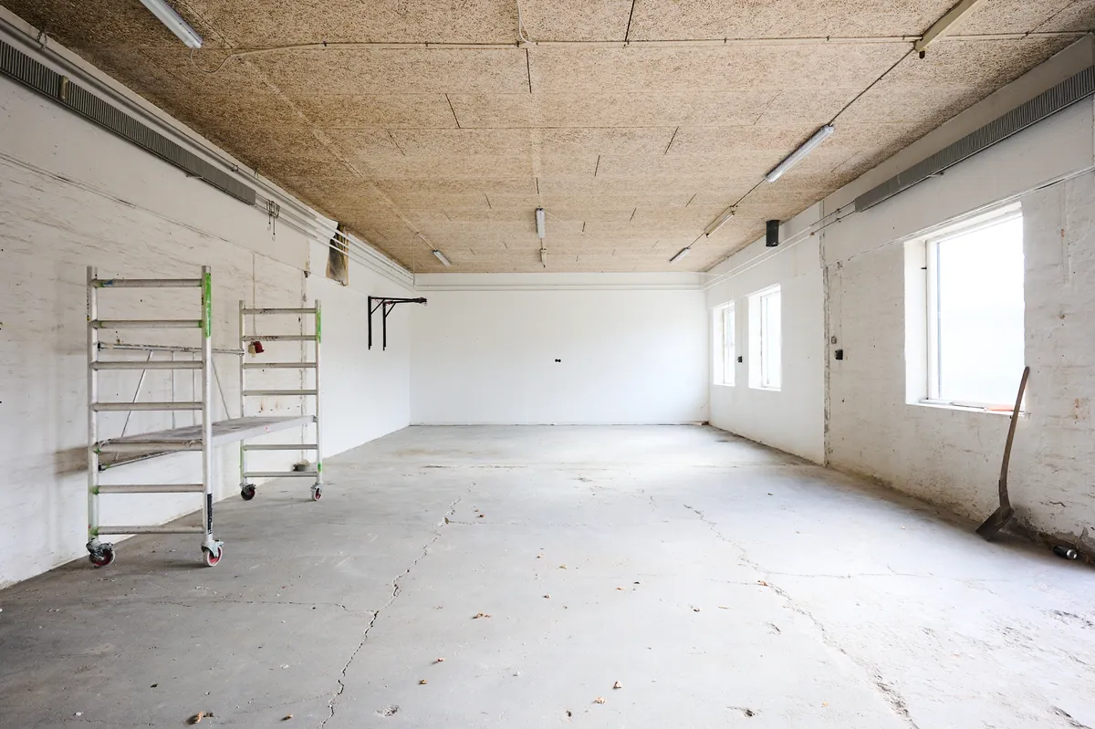 Betongulv med spor af skrald og revner, højt loft med lysekroner, to skraldspande, stående stige, to vinduer og jordskovlen