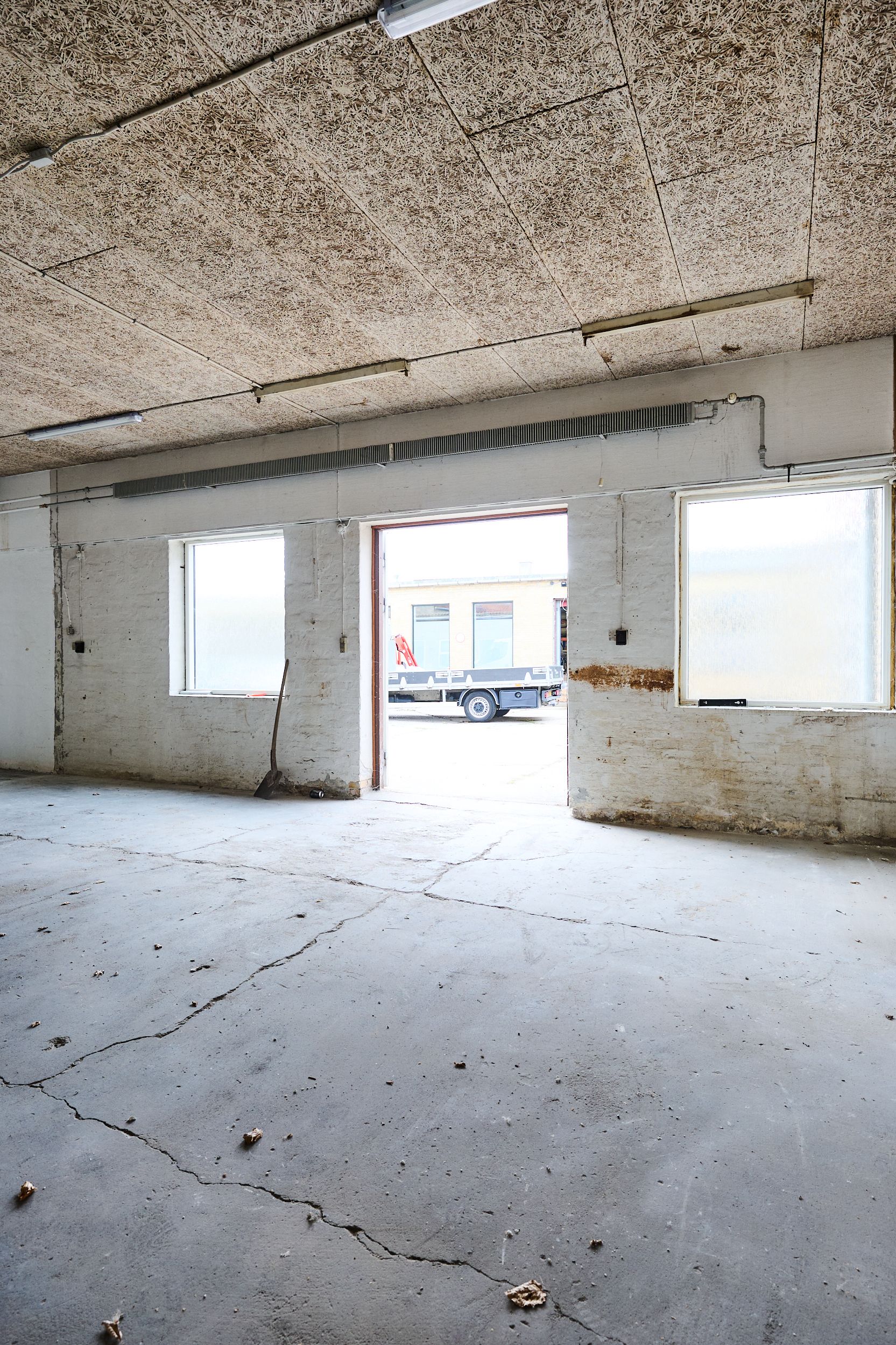 En stor betongulv, skrald og et værktøj. Vindue og åben dør med fragtvogn. Lys på loftet og rørklædt væg.