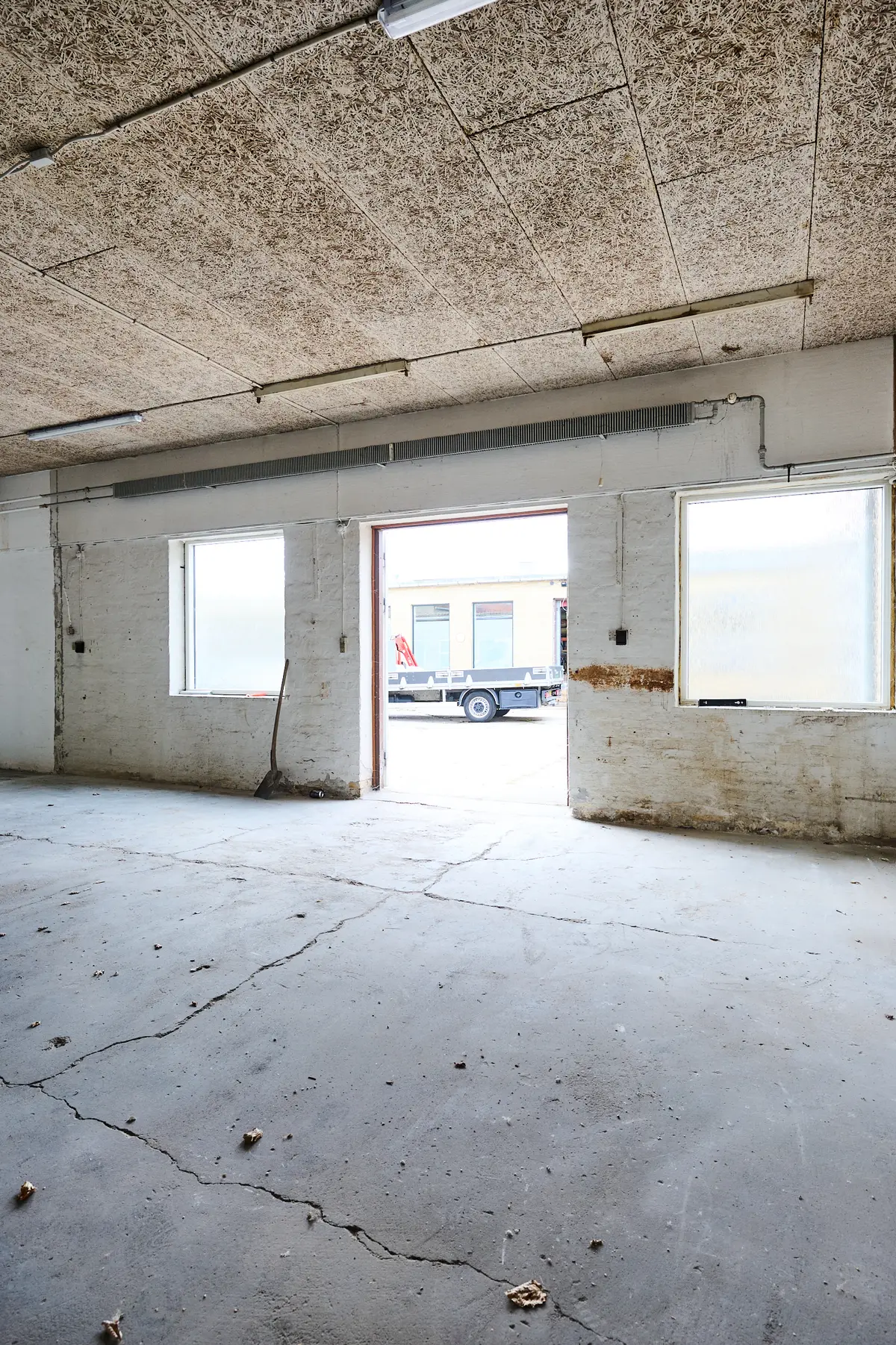 En stor betongulv, skrald og et værktøj. Vindue og åben dør med fragtvogn. Lys på loftet og rørklædt væg.