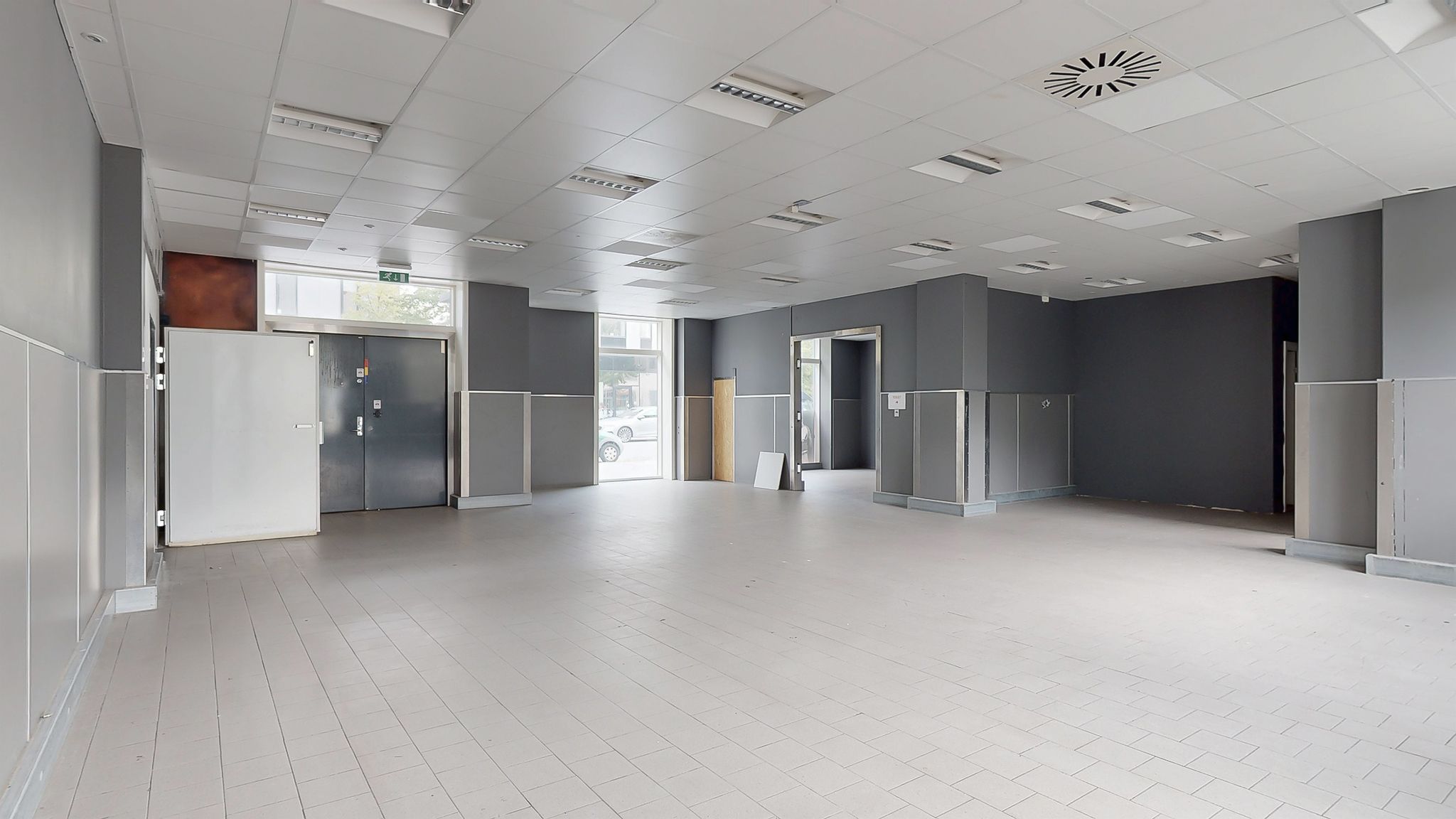 Et tomt lokale med hvide vægge og gulv, højt hvidt loft, glasvinduer og to døre i hver ende af lokalet