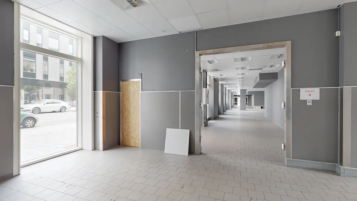 Lang, tyst gang mellem to vægge, en dør åben og en toiletskilt, gulvet og loftet er hvide, og der er skarpe skygger