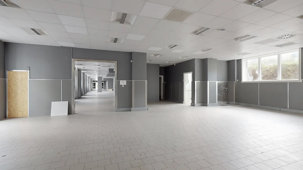 En stor, tom gang med hvide gulvfliser og grå vægge, hvor vinduer og døre er lukket. Væggene er uden møbler og udsmykning