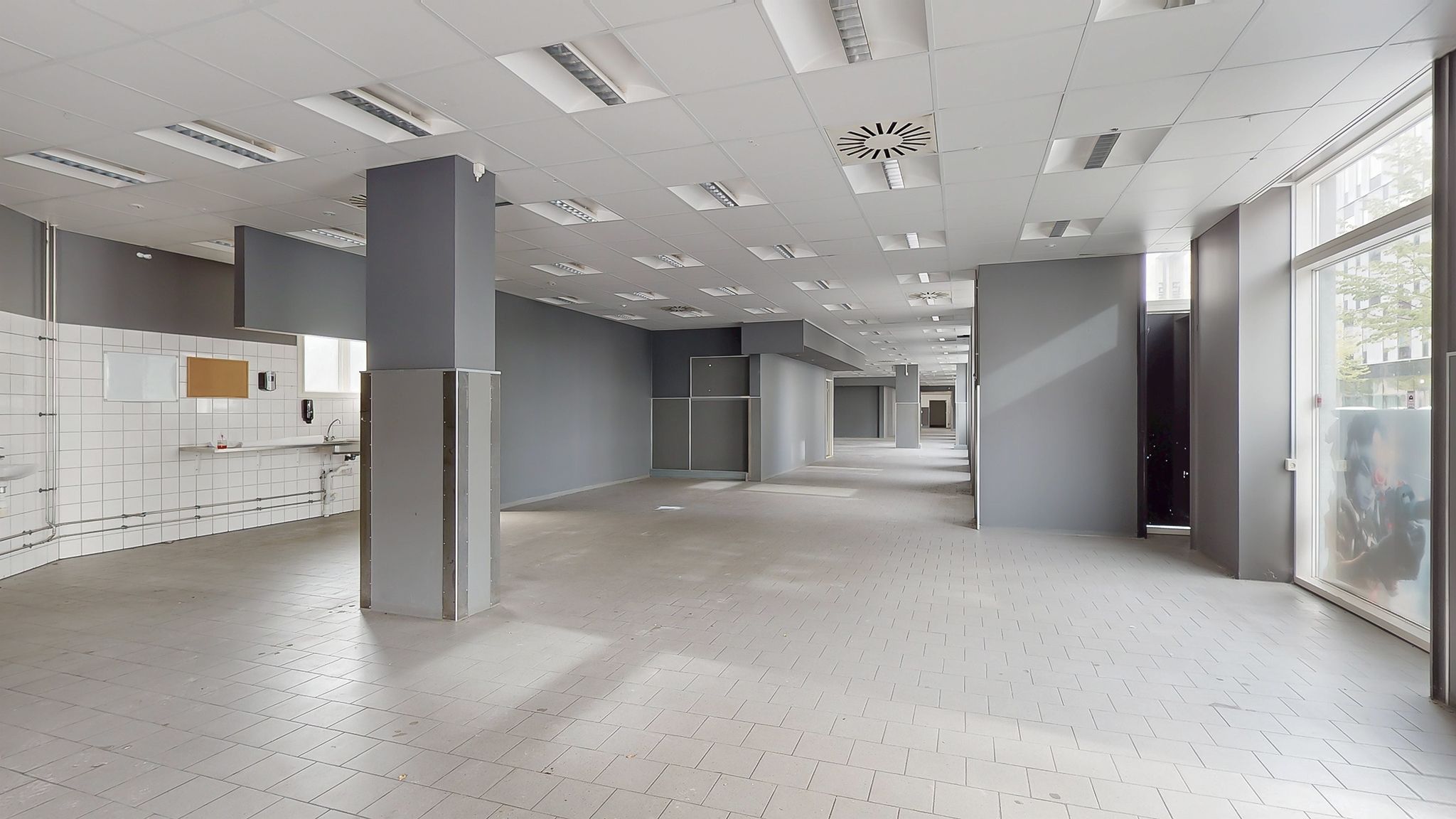 Lang og tom gang med hvide kakel og lyse gulvbelægning. Moderne ventilation og lysekroner på loftet. To større søjler i