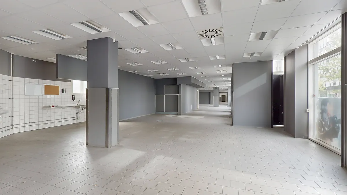 Lang og tom gang med hvide kakel og lyse gulvbelægning. Moderne ventilation og lysekroner på loftet. To større søjler i