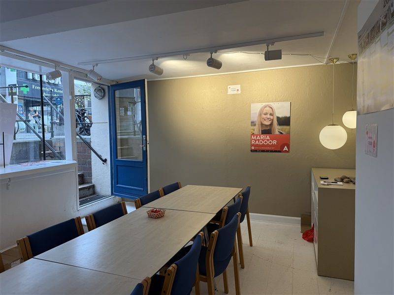 En rummelig kontorlokal med lange borde og blå stole, Maria Radoods navn og billede på væggen og en lysekroner hængende på