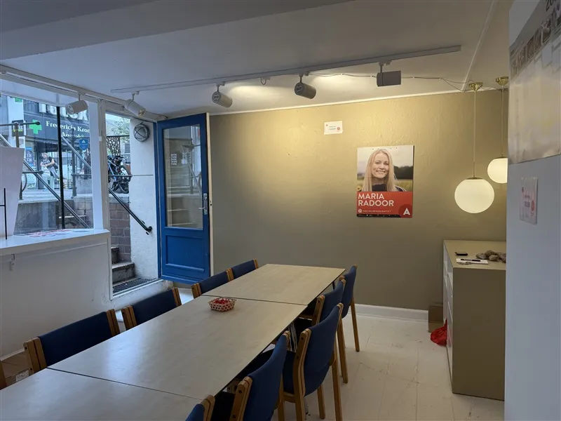 En rummelig kontorlokal med lange borde og blå stole, Maria Radoods navn og billede på væggen og en lysekroner hængende på