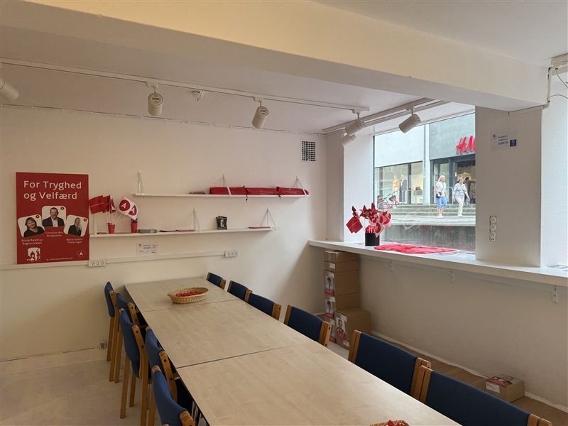 Inde i lokale med lyse vægge og lofter er der borde og stole samt en vinduesvisning med mennesker udenfor