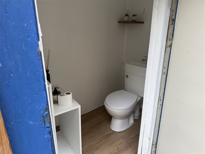 En lille toiletrum med en hvid toilet med lukkedes og et hvidt hylde med toiletpapir og en pumpeflaske.