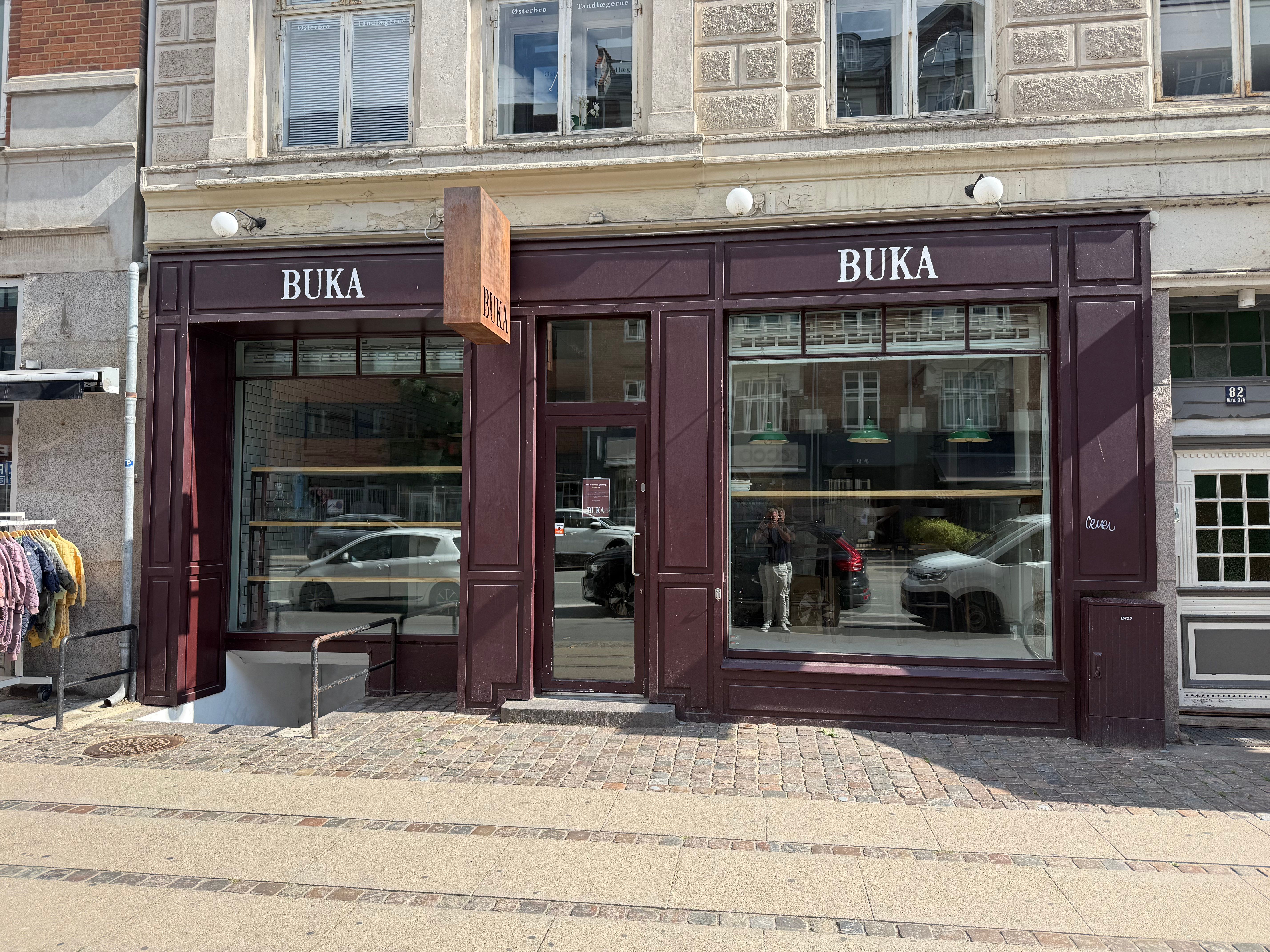 Indgående forretningslokaler med to vinduer og en dør i rødt. Indersiden viser flere produkter og et par mennesker. Væggene