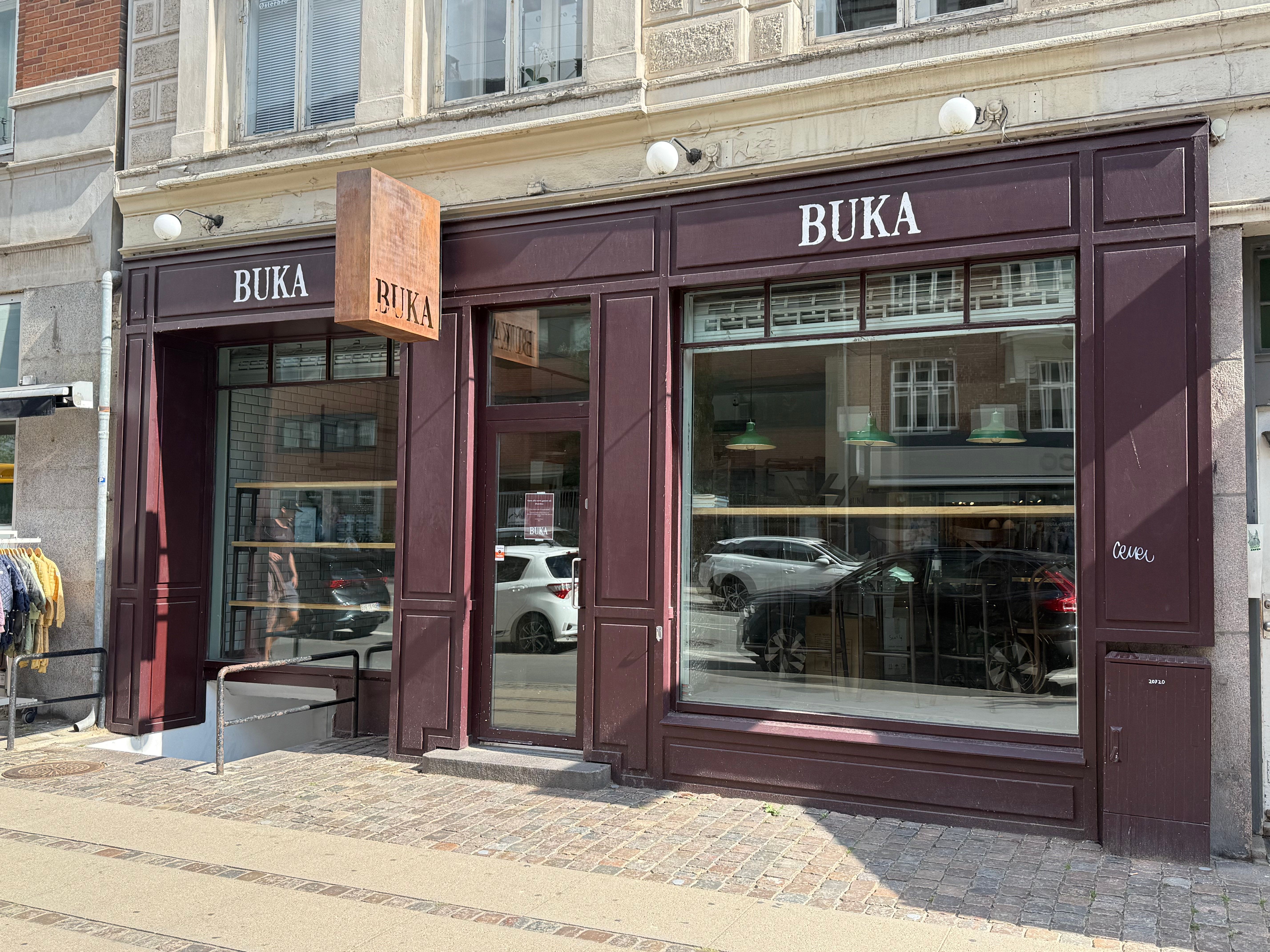 En elegant boutique, Buka, med glasdøre og vinduer reflekterer biler og en kvinde i baggrunden, hvor tøjvægge og lysekroner