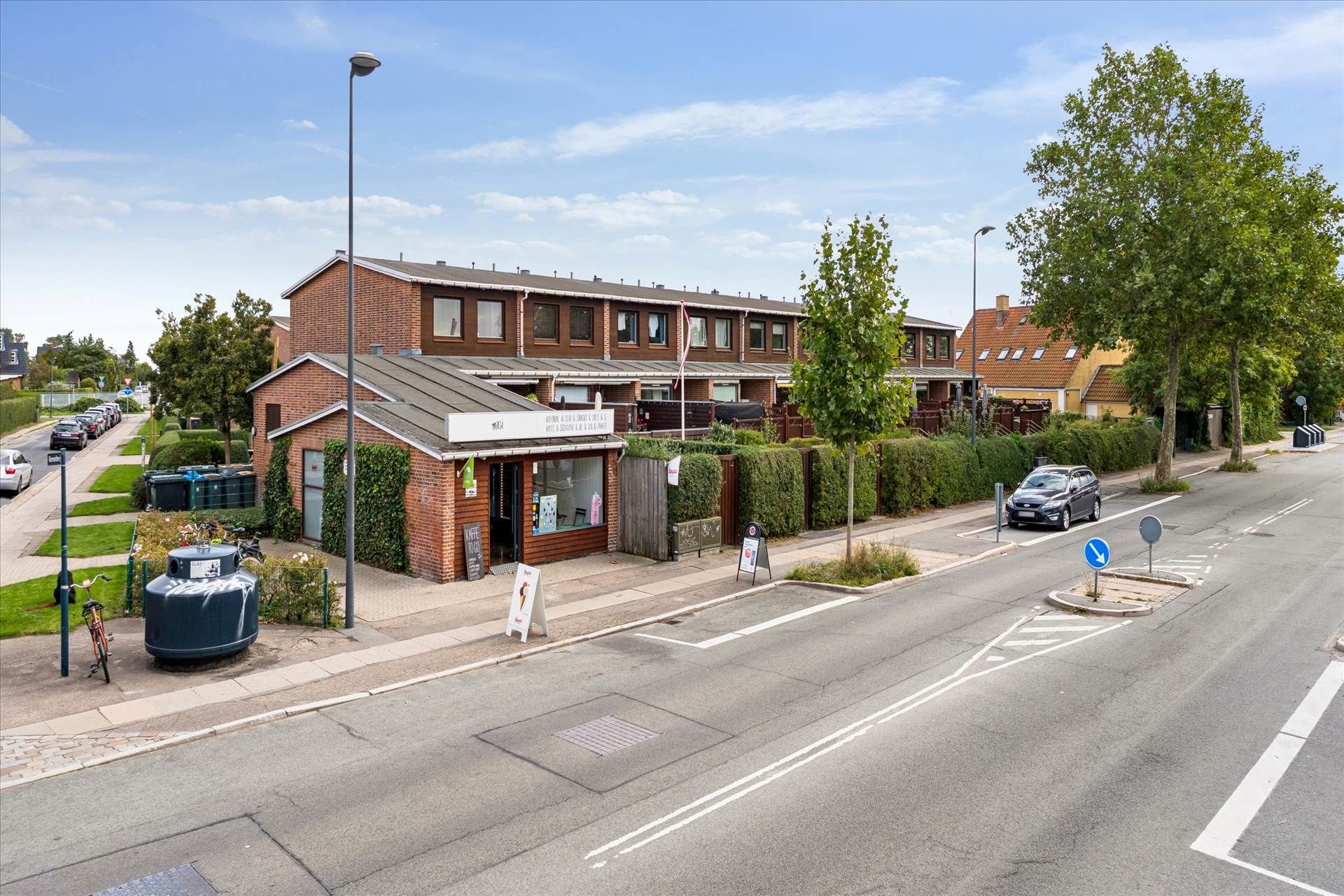 Kiosk med skilt og terrasse, foran parkeringsplads med cykel og søjle. En bil kører på den tosporede vej, der er mærket med