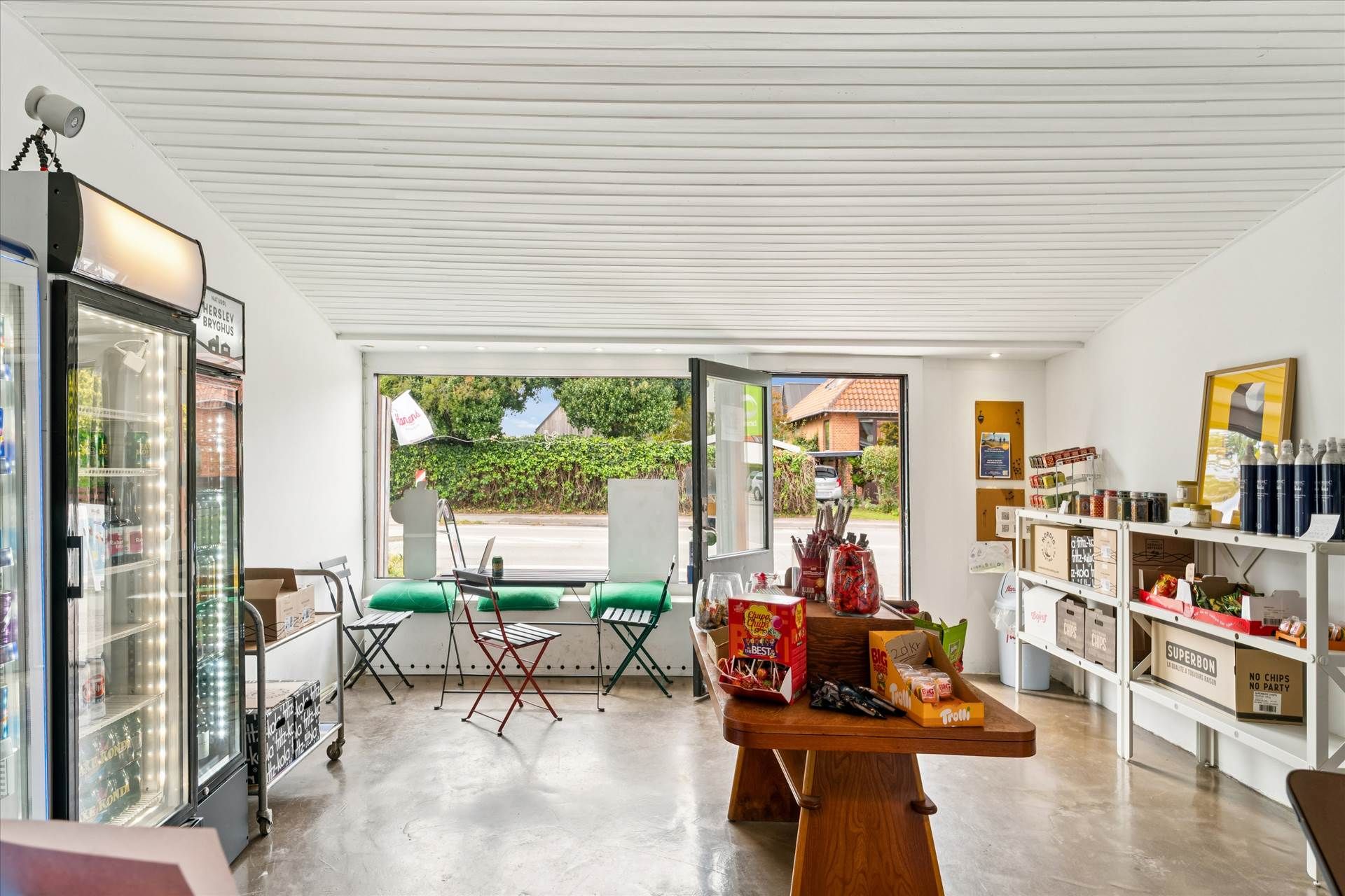 En kiosk med en stor glasdør og vinduer, indeholder køleskabe med snacks og drikkevarer og en bord med sæder.