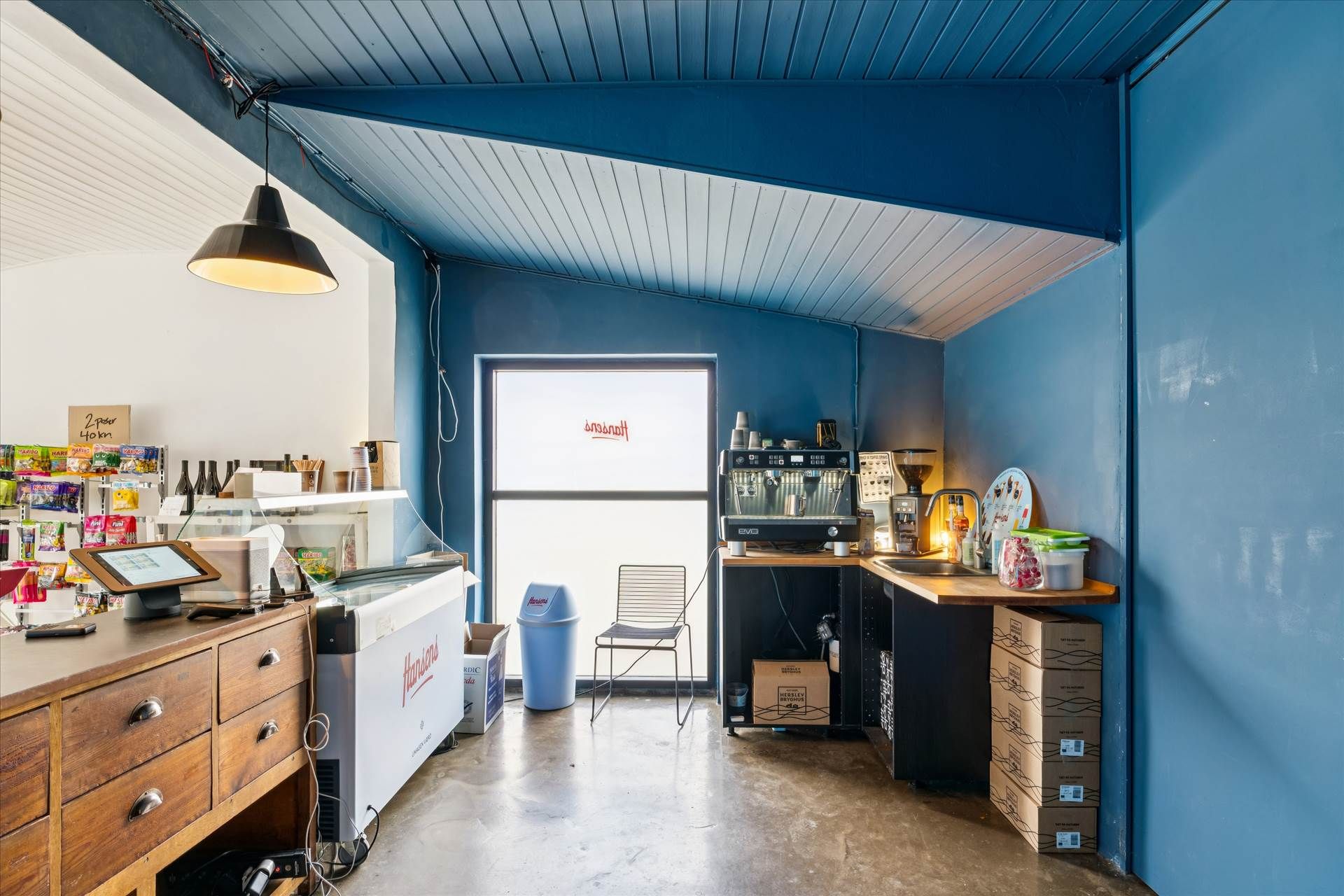 Kiosk med indretning og møbler, herunder en stol, et køleskab, en espresokaffemaskine, en skraldespande og en skrivebord med