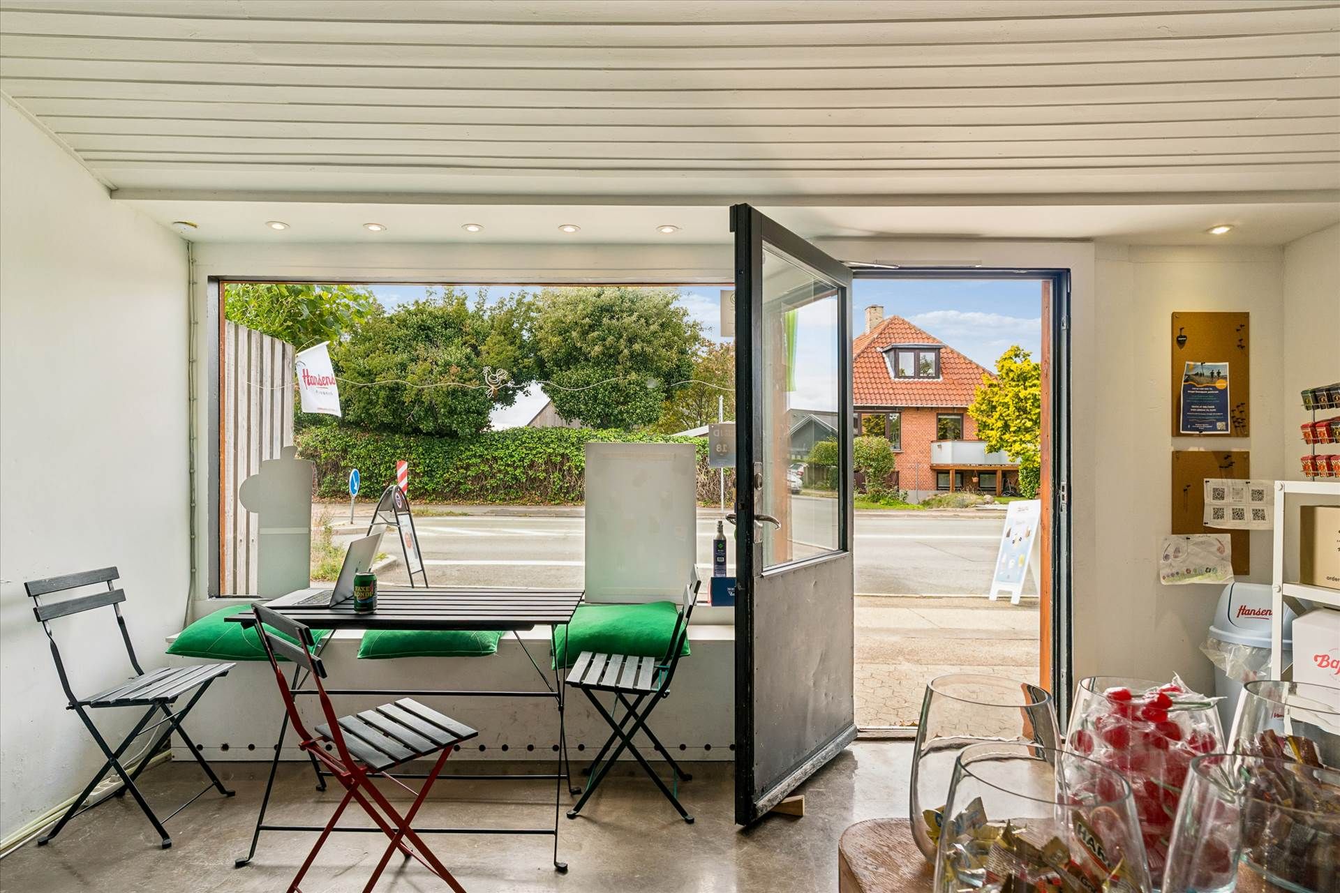 Kiosk med åben dør og udsigt til vej og bygninger, borde og stole samt slik på hylder og i glas