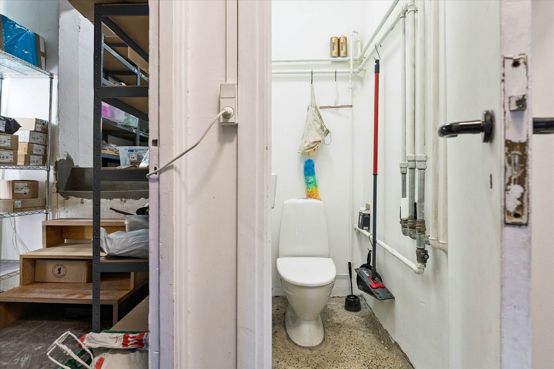 En toiletrum med en hvid toilet, en børste og en spidset skovl i et lille rum med mange rør og en dør til en lagerrum
