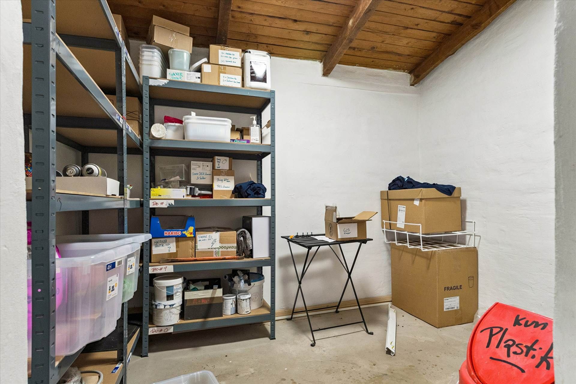 Lagerrum med stelhylder og bokse med diverse ting, bl.a. spraydåser, plastbeholdere, malerhinker og en gul bænk.