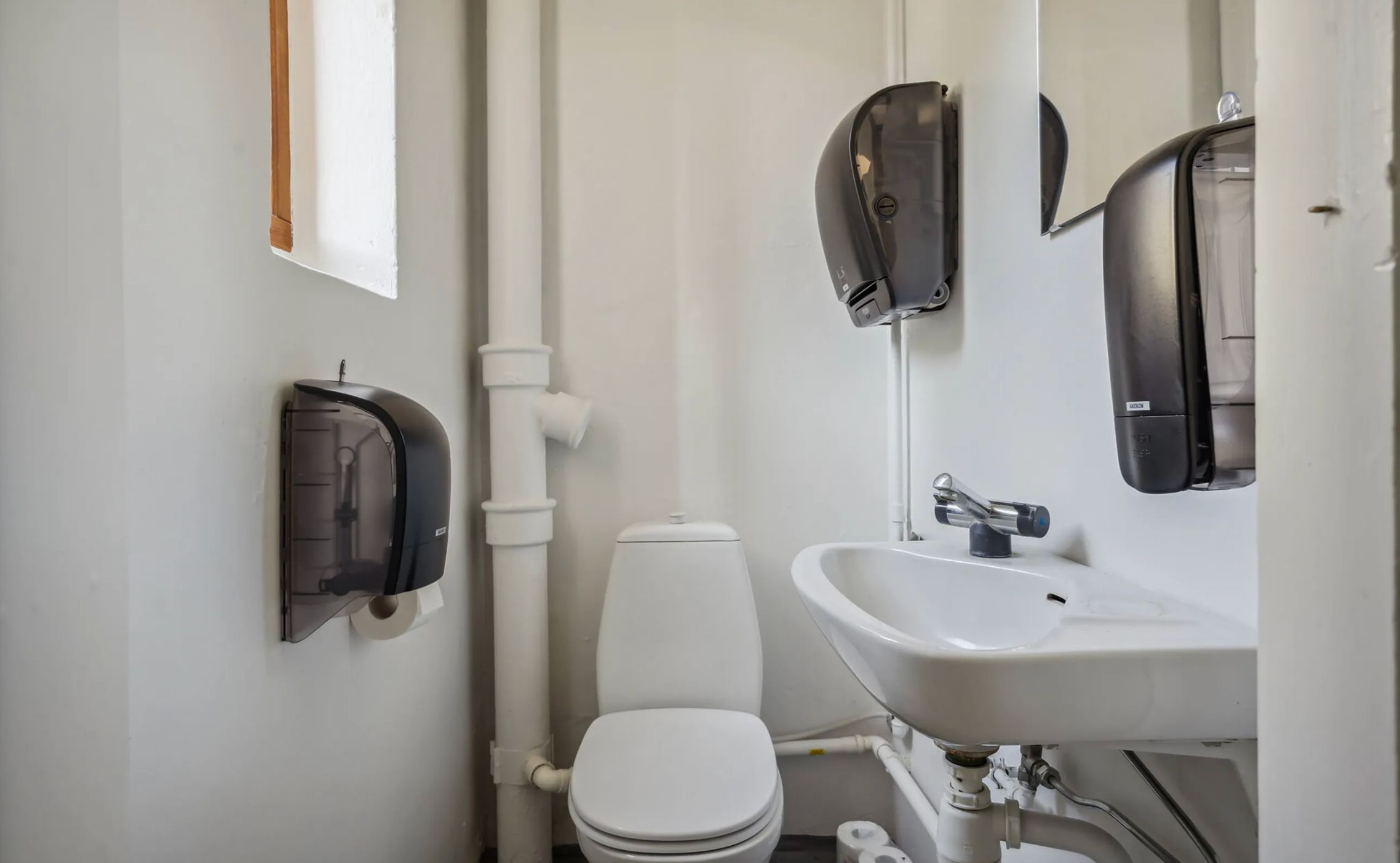 Toilette med toiletpapirautomat, hvid vask og spejl med håndklædeautomat i et hvidt lokale