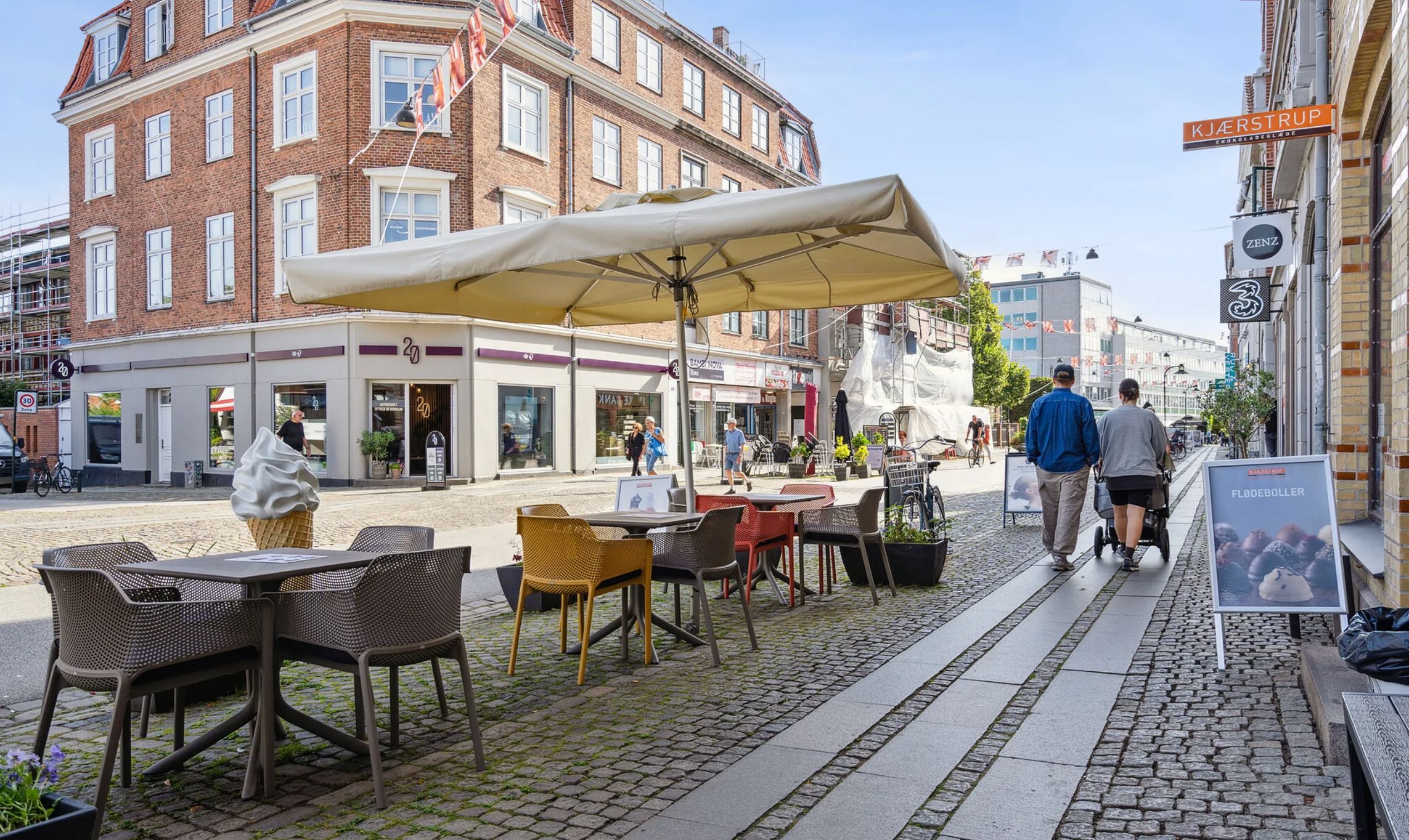 Mennesker går i gågaden, der er fyldt med planter og borde med stole samt en is med paraply på denne solrige dag