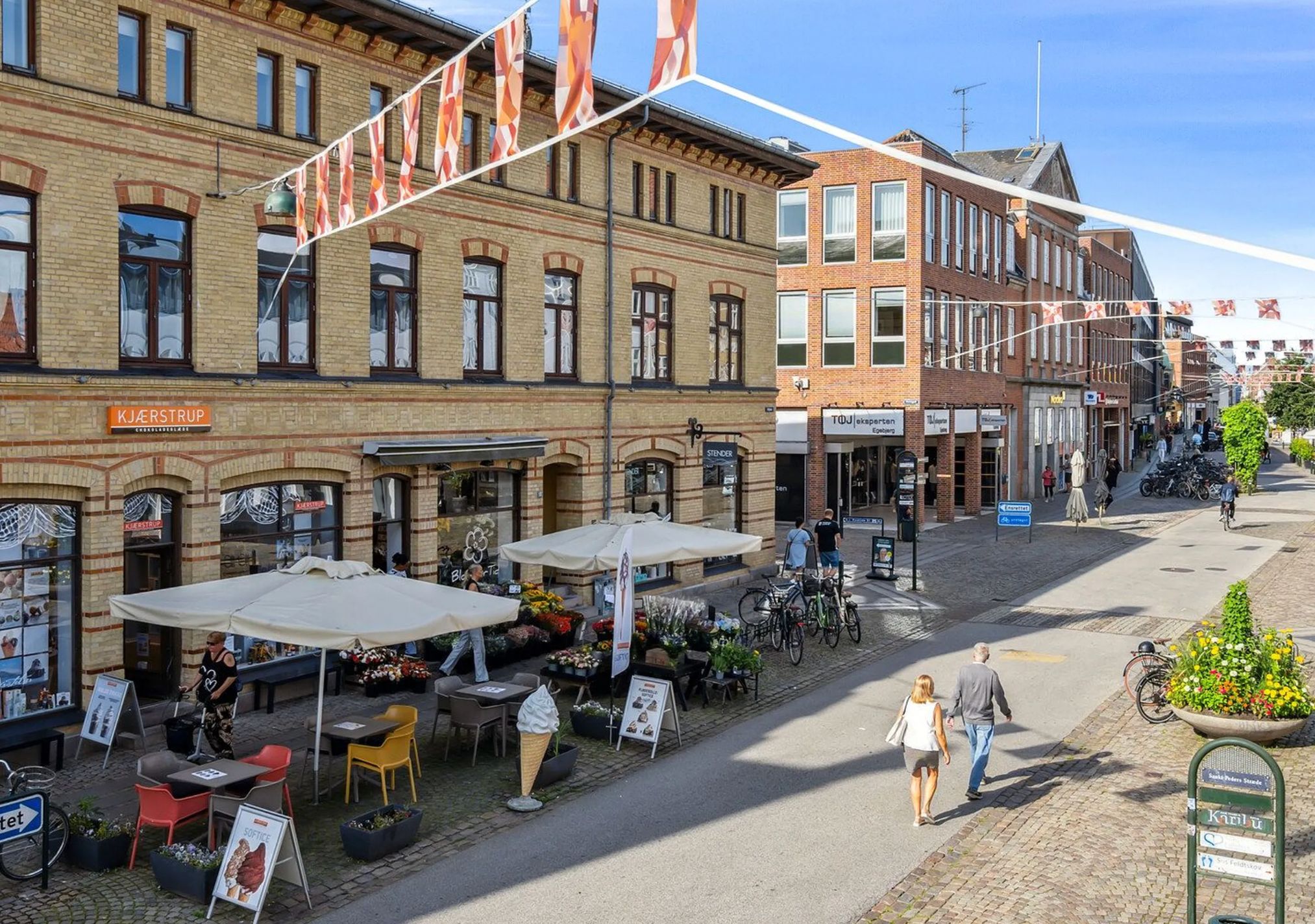 Gågaden i Roskilde med blomsterstande, parasoller, iskrukker, cykler og mennesker der går forbi med poser