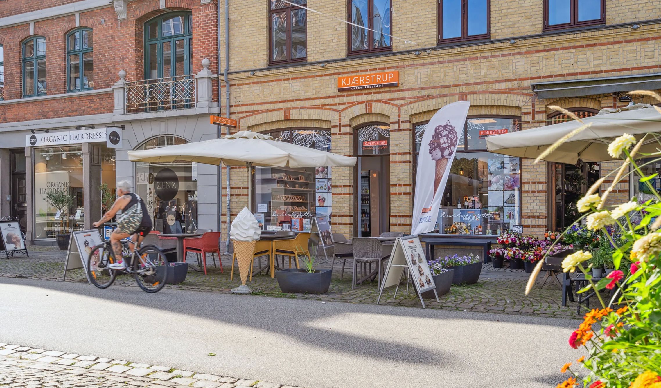 En cykelkørende mand kører forbi en gade med blomster og forretninger med cafeer og butikker i Roskilde