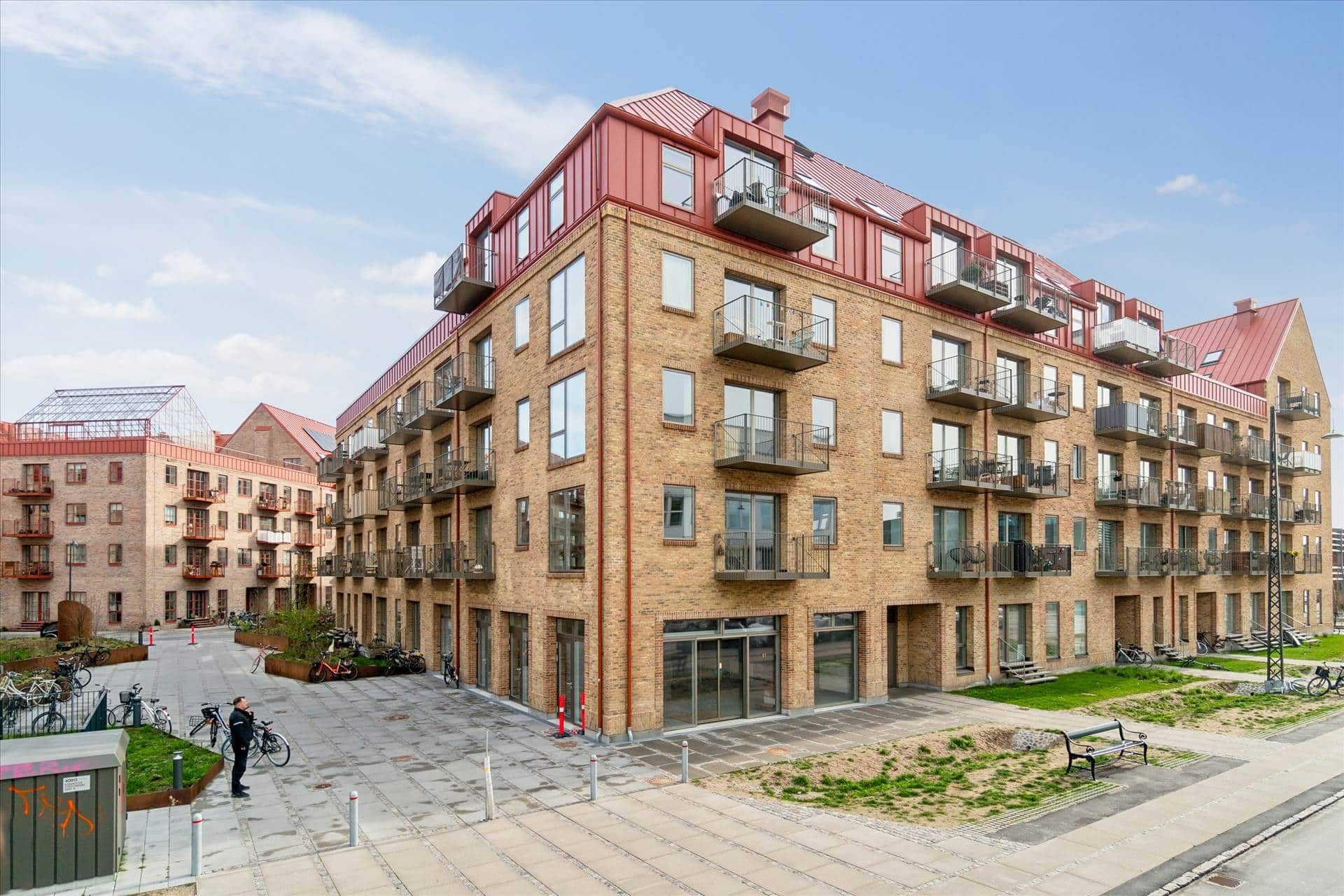 Moderne lejlighedsbygning med rød tagbeklædning og terrasser i København, parkeringsplads med fikserede cykler og grønne