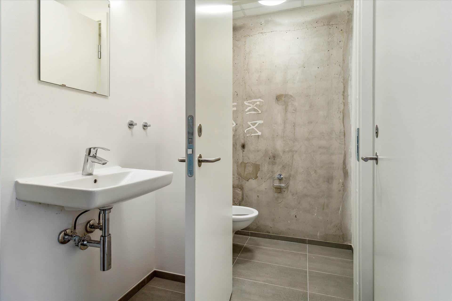 Bad med hjørnebadekar, toilet, håndvask og spejl. Lys og moderne interiør med lysegule fliser og lysegrå vægge.