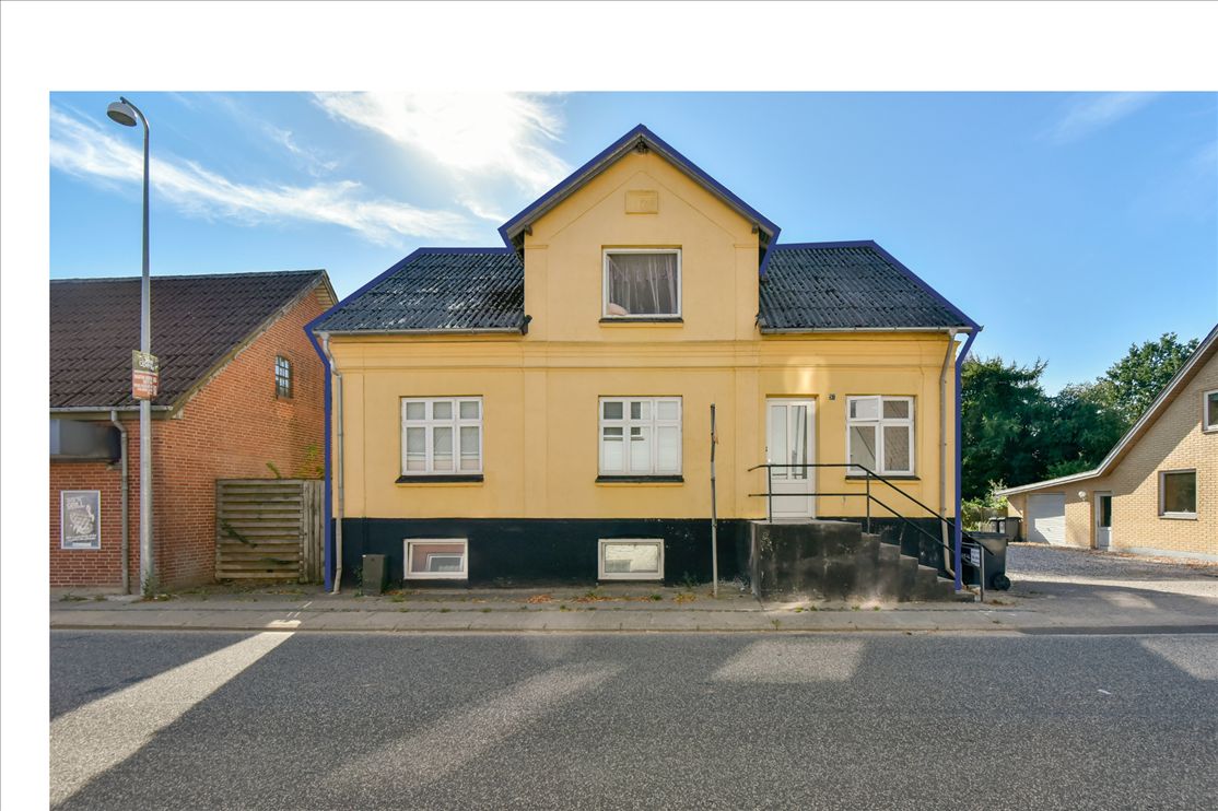 Gade med solbelyst, gult hus med tegltag og hvide ruder og grønne træer på baggrunden