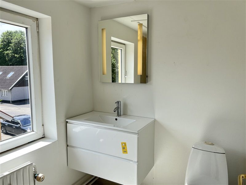 Et badeværelse med et hvidt toilet, en hvid badeværelsesskab og en spejl med lyse gule kant. Et køretøj udenfor og træer i