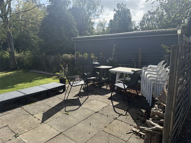 Velholdt terrasse med udendørs møbler og et bord samt træbænk og en række hvide stole på baggrund af grønne planter og træer