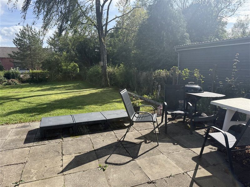 Terrasse med træstole og bord, liggestol, græsplæne, buske og træer i baggrunden, og solskin lyser igennem trækronerne