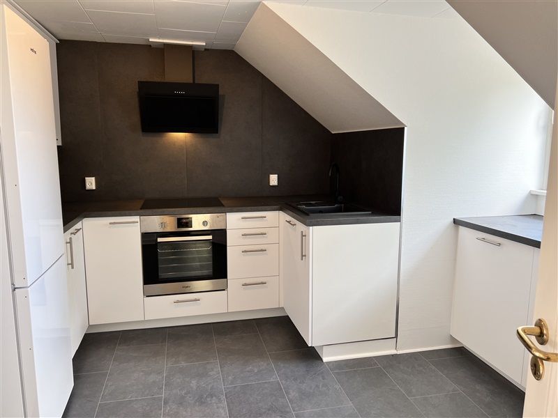 Velholdt køkken med hvide skabsfronter og grå gulv. Sort elektrisk komfur, indbygget ovn og lysegråt loft.