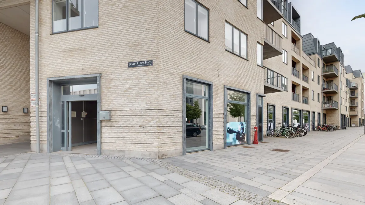 Bygning med gavl og glasdøre, med skilt for Jesper Kleins Plads. Biler og cykler parkeret foran og facade med lysebrune