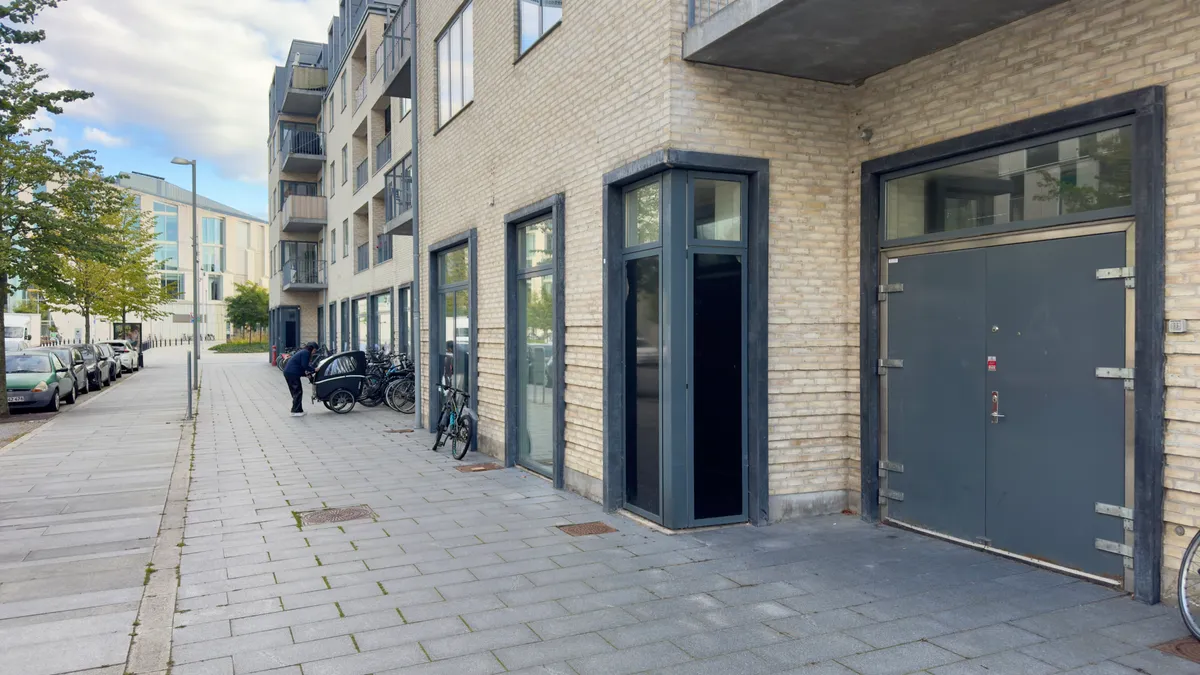 En mand lægger et cykelbarnevogn på en brolægget gågade med en bygning i baggrunden og flere parkerede biler og cykler