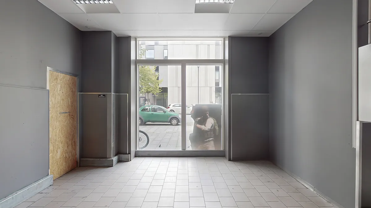 Et tomt rum med hvide gulvfliser og grå vægge, en åben dør og et glasdørfælde med refleksioner af biler og en person i