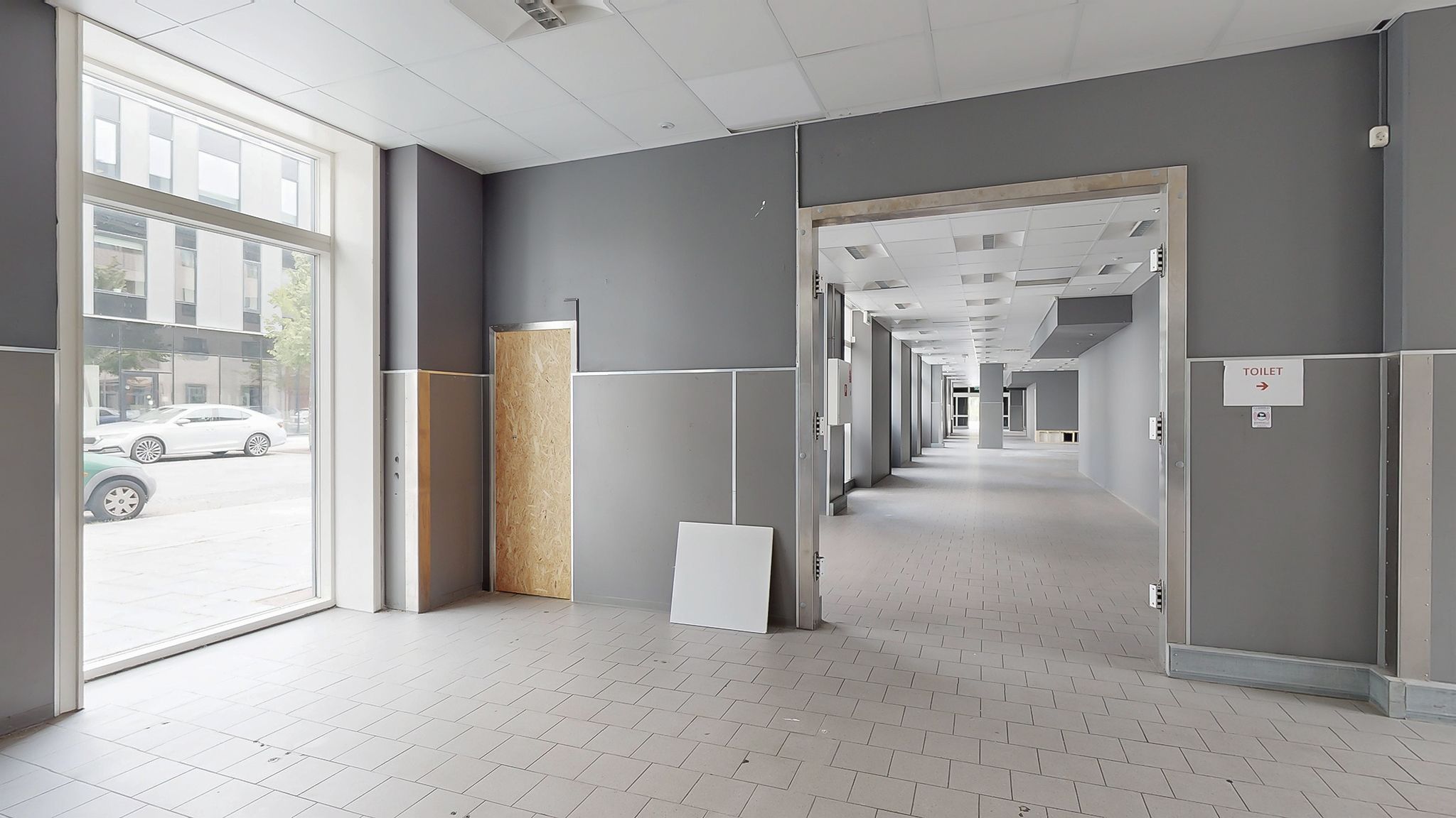 Langt korridor med toiletskilt og to åbne døre, en dør med forsiden af træ og vinduer med udsigt til en bygning og biler på