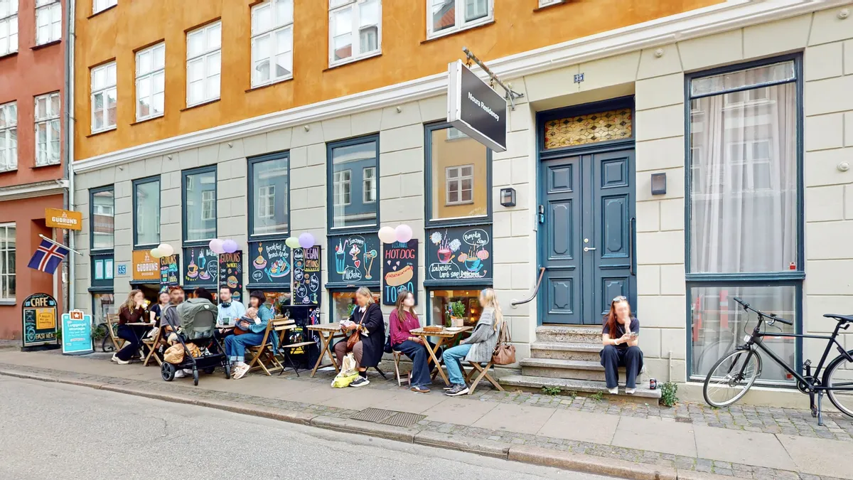 Kvinder og mænd nyder morgenmad og hotdogs udenfor caféen Nouras Residence i København med en cykel og en norsk flag ved
