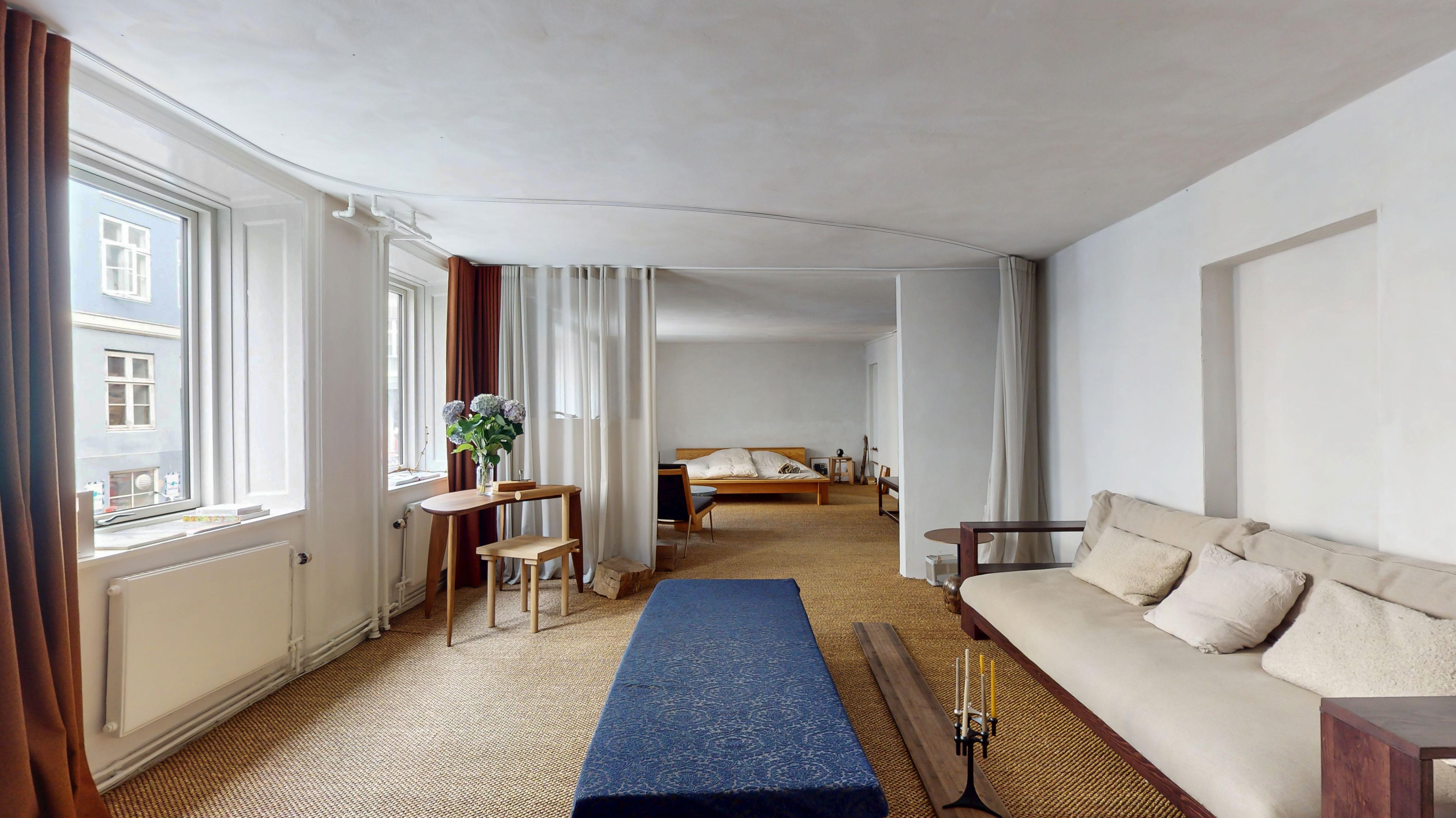 Minimalistisk stue med lyse vægge, hvidt loft og gulvtæppe, sofa med puder, bord og stol, vinduer med gardiner og møbler i