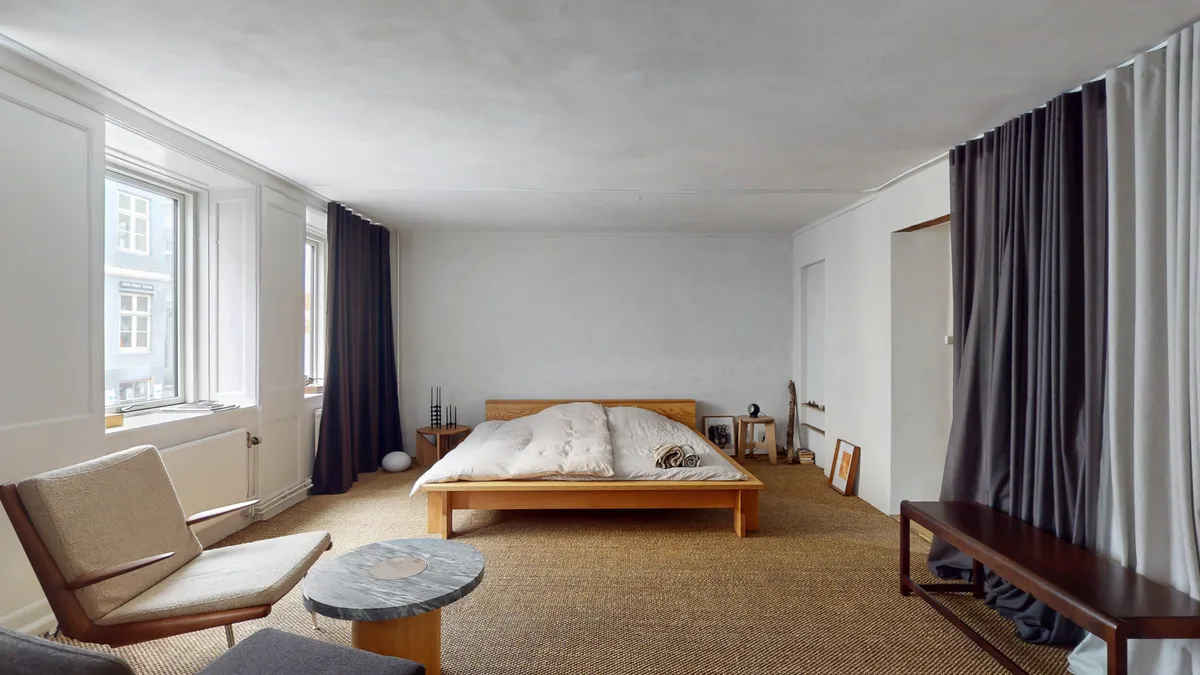 Et moderne soveværelse med en hvid væg og en trægulvved. Et enkelt træseng med hvide dynehoveder og et træbord med en runde